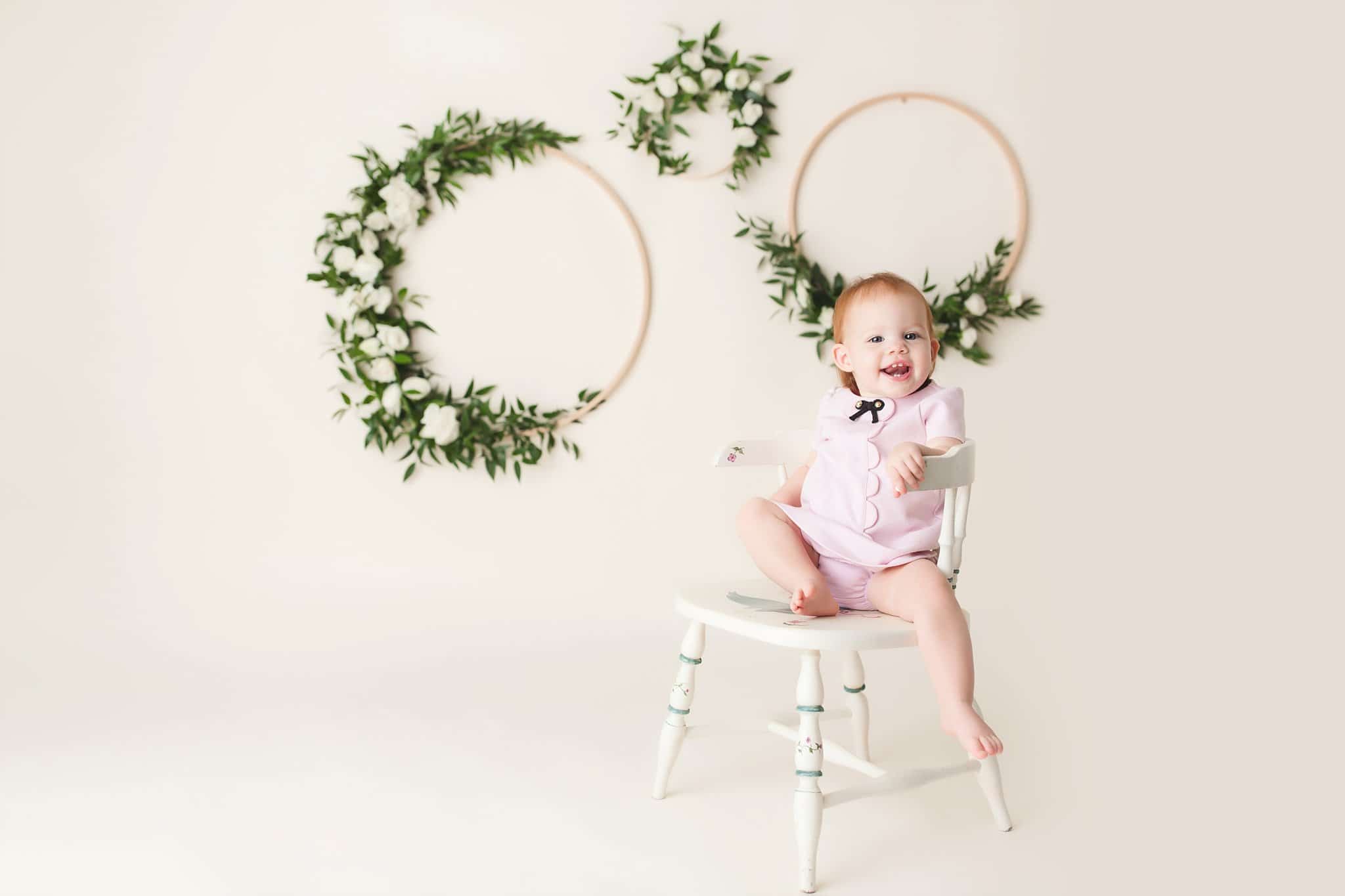 toddler girl sitting white chair cream background floral wreaths 1st Birthday Pics Jacksonville