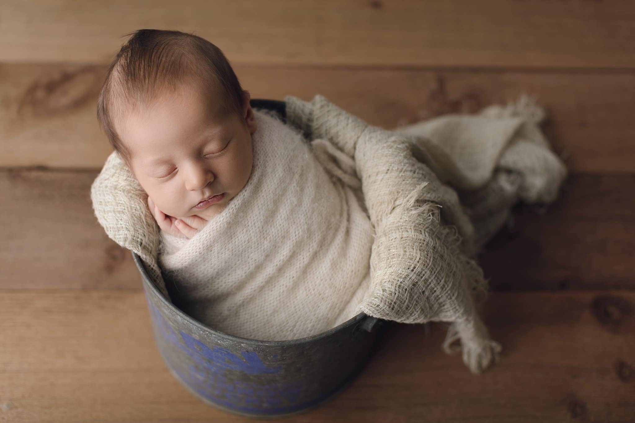 newborn baby boy swaddled in cream and sleeping in vintage bait bucket on wood background