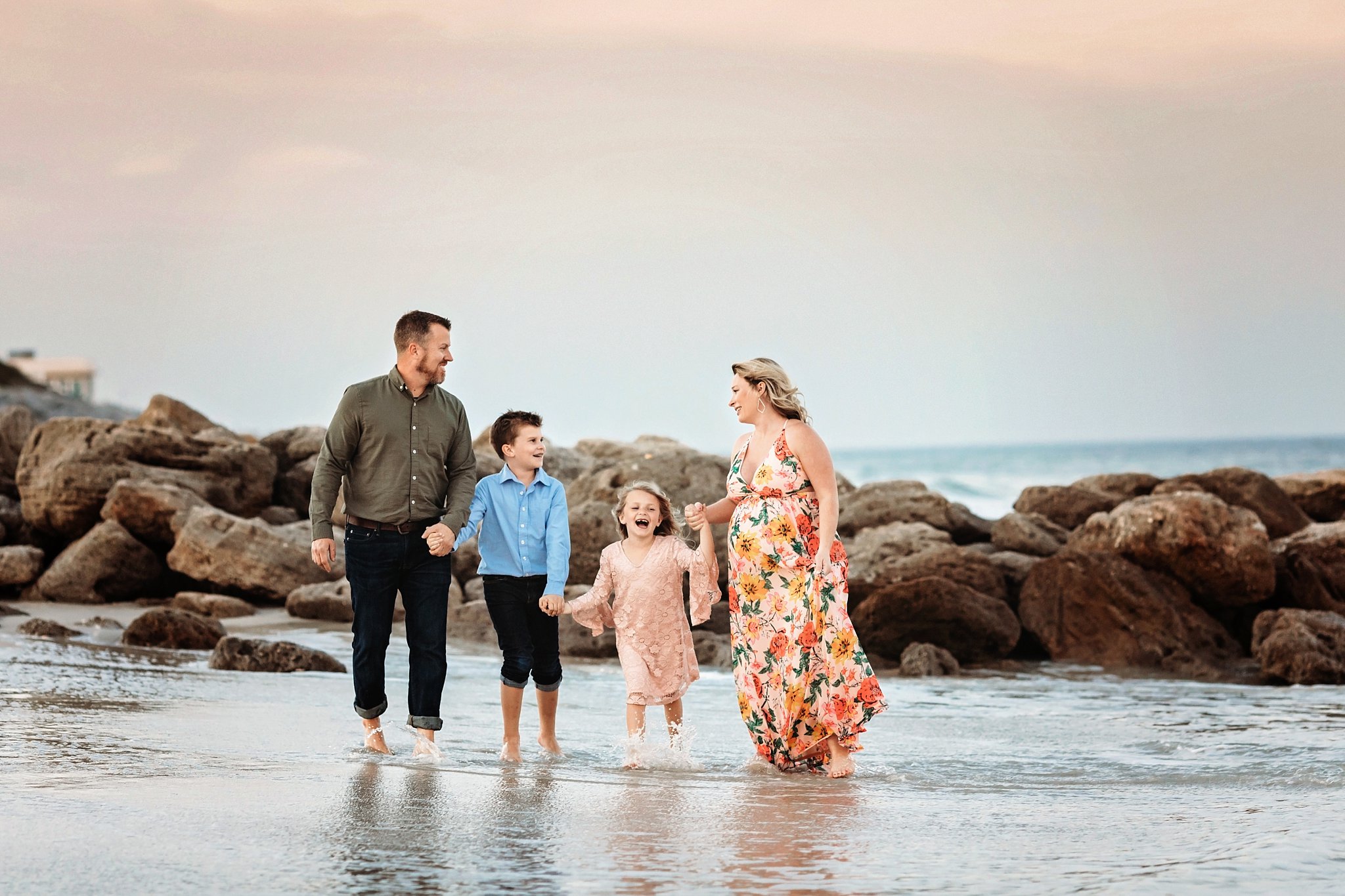 St Augustine Maternity Photographer Washington Oaks Gardens State Park family of 4 walking in the waves with pink sky and rocky beacg behind them