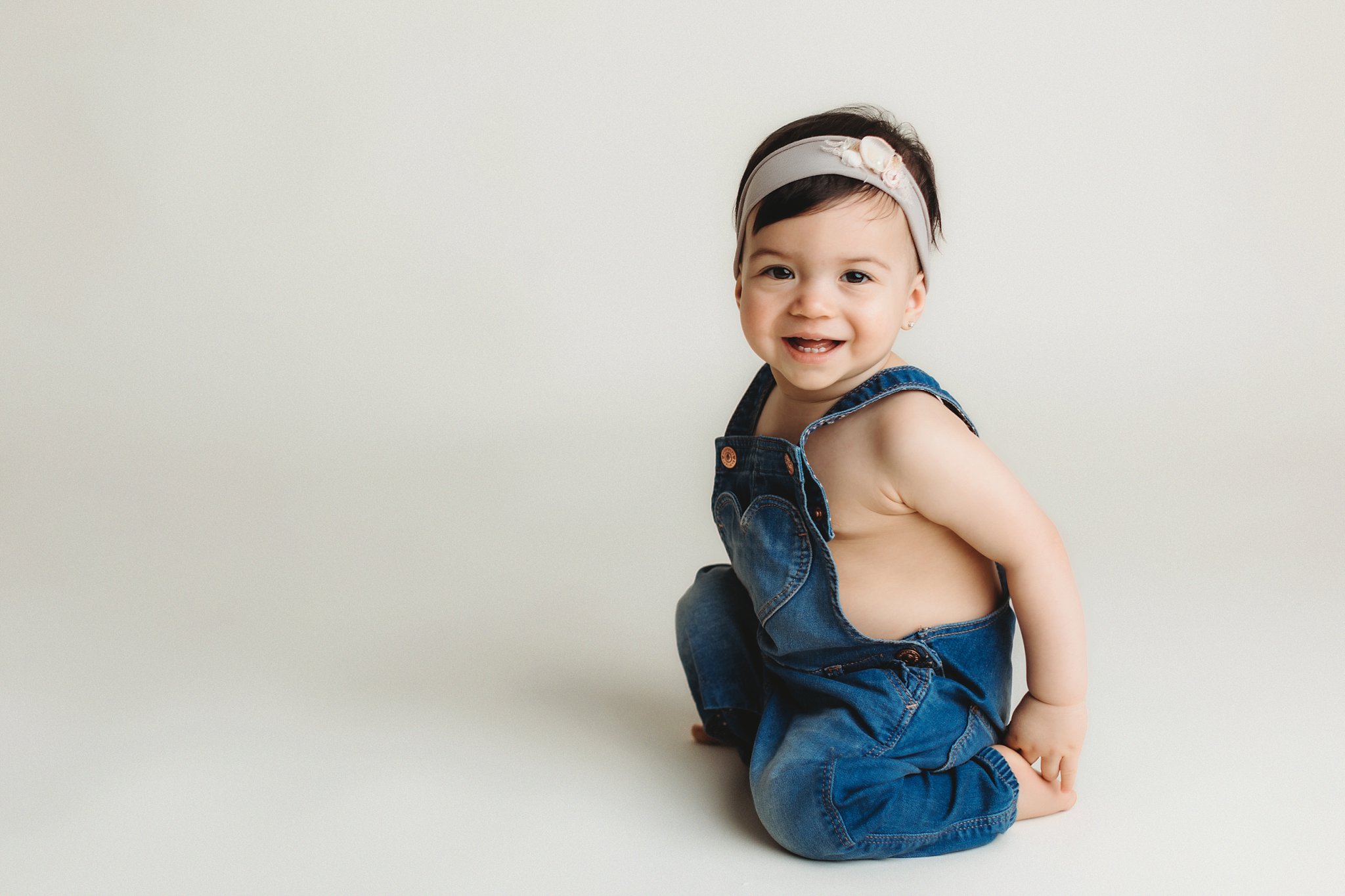 1 year old little girl on cream seamless background in denim overalls and pretty headband