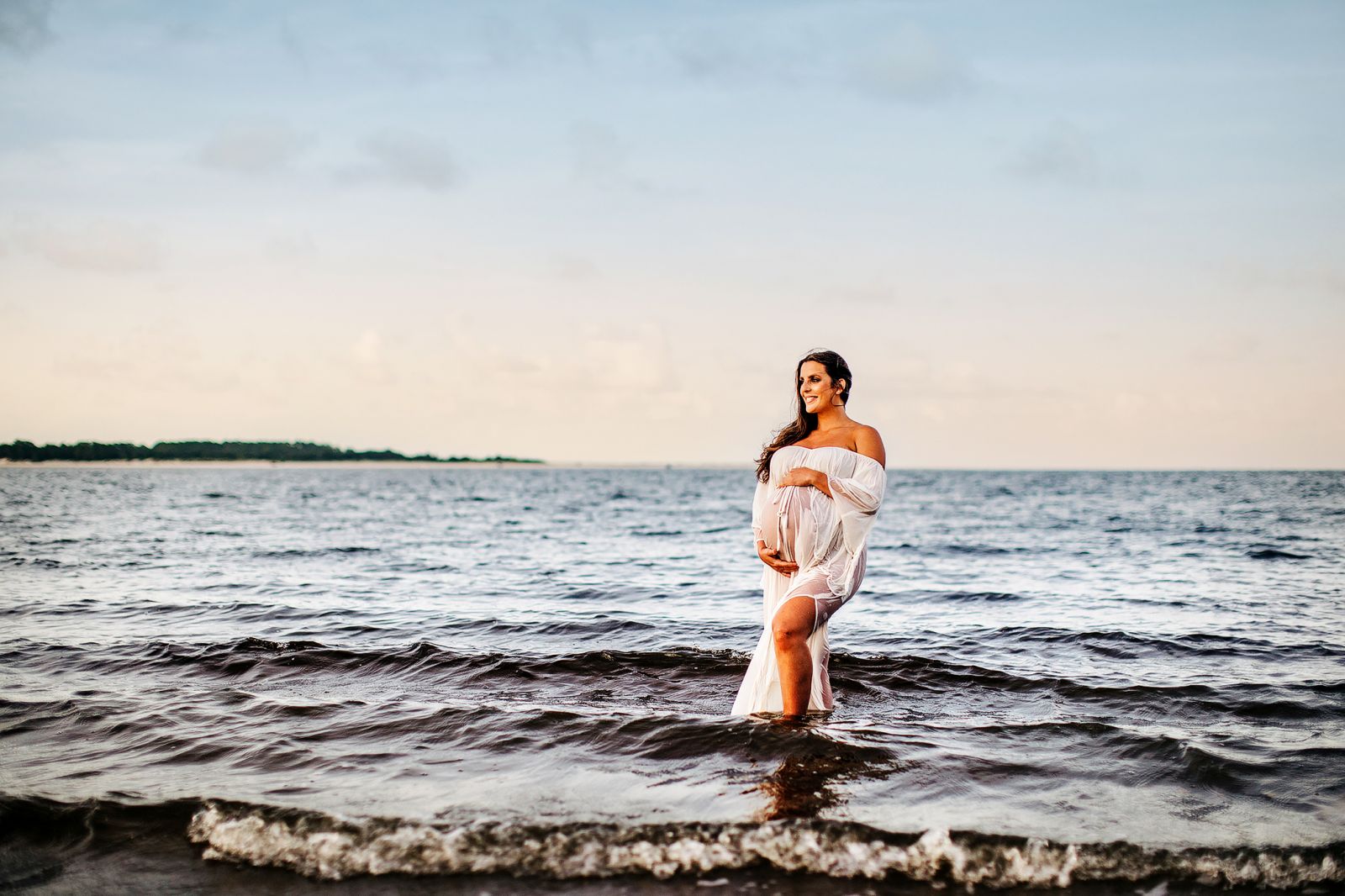 maternity photo of mom with long dark hair in sheer white gown standing in the ocean