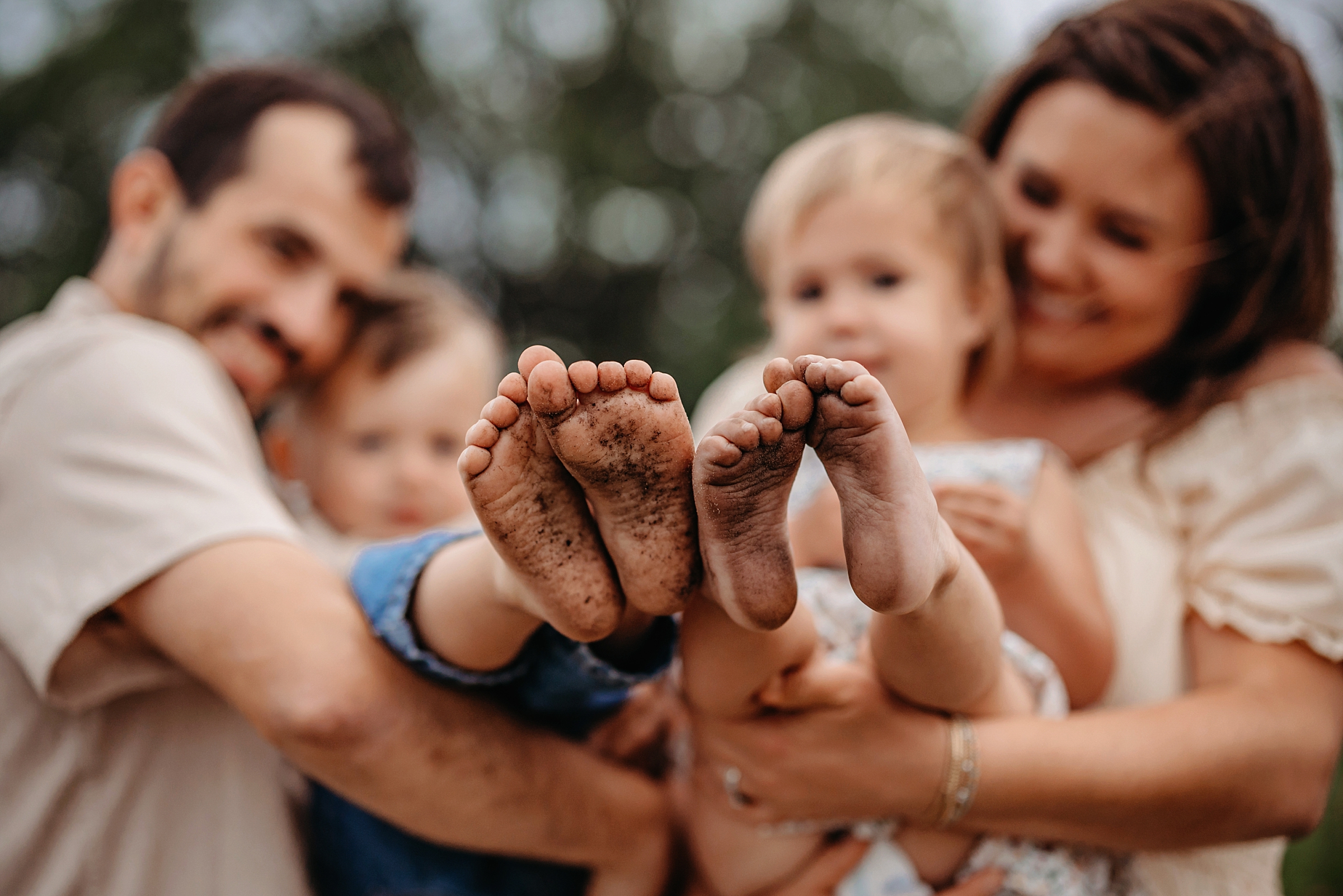 Close-up of two children’s muddy bare feet with their smiling family softly blurred in the background.