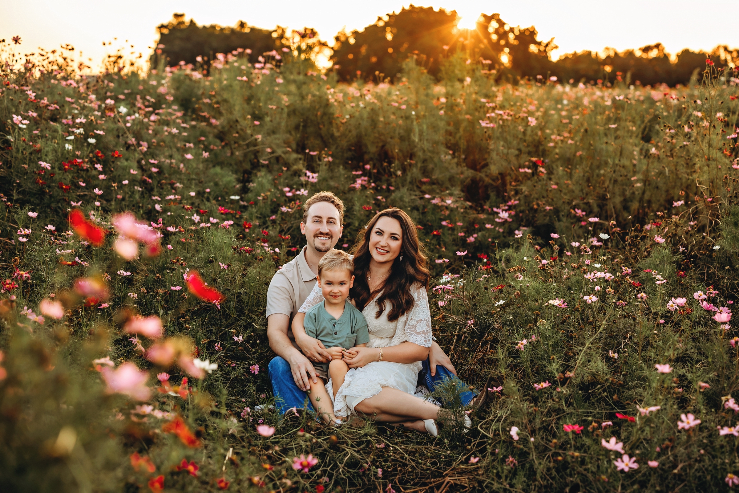 Family of three sitting together surrounded by tall pink wildflowers with the setting sun behind them.