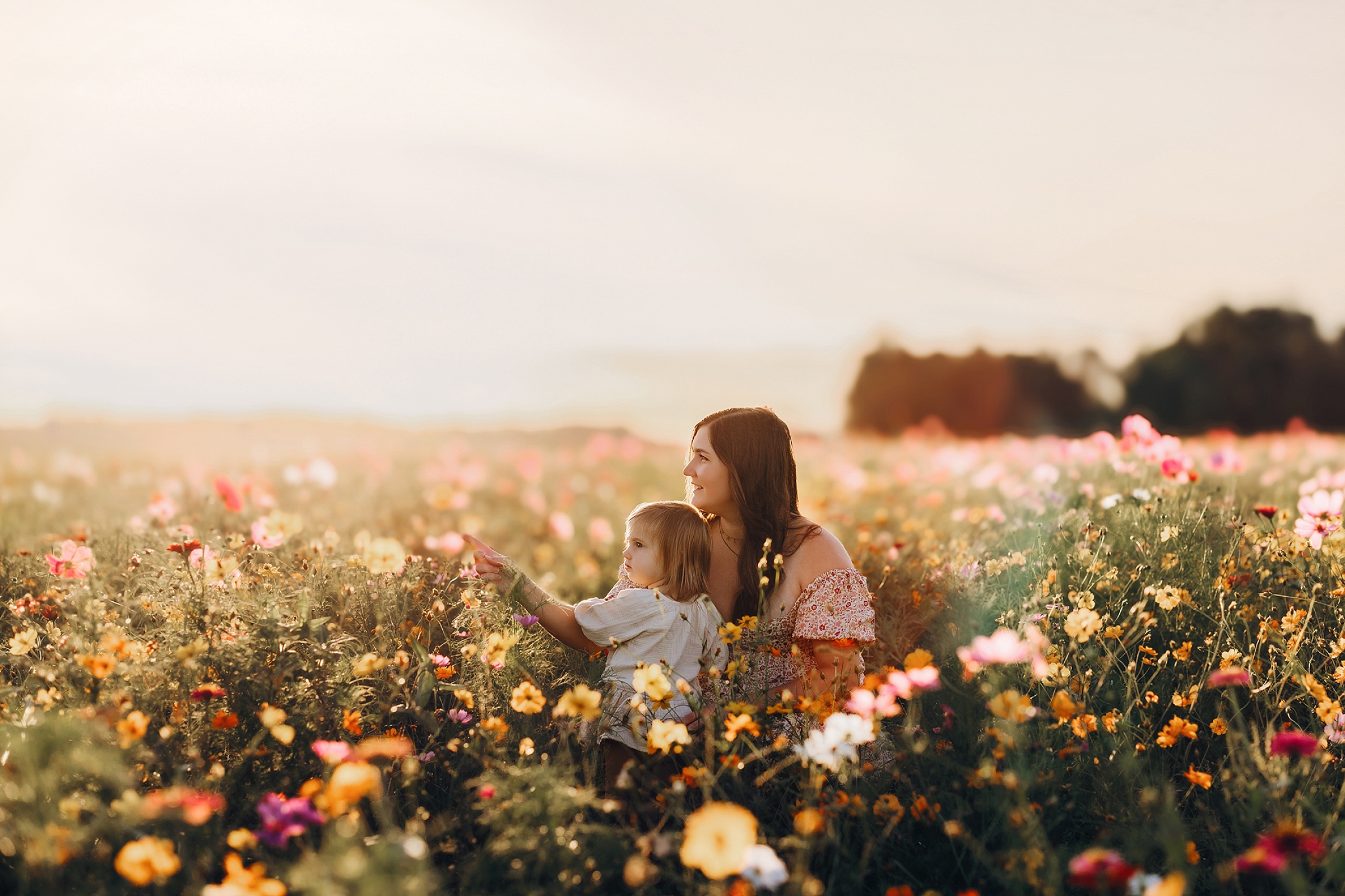 Mother and young daughter sitting together among colorful wildflowers in warm golden evening light.