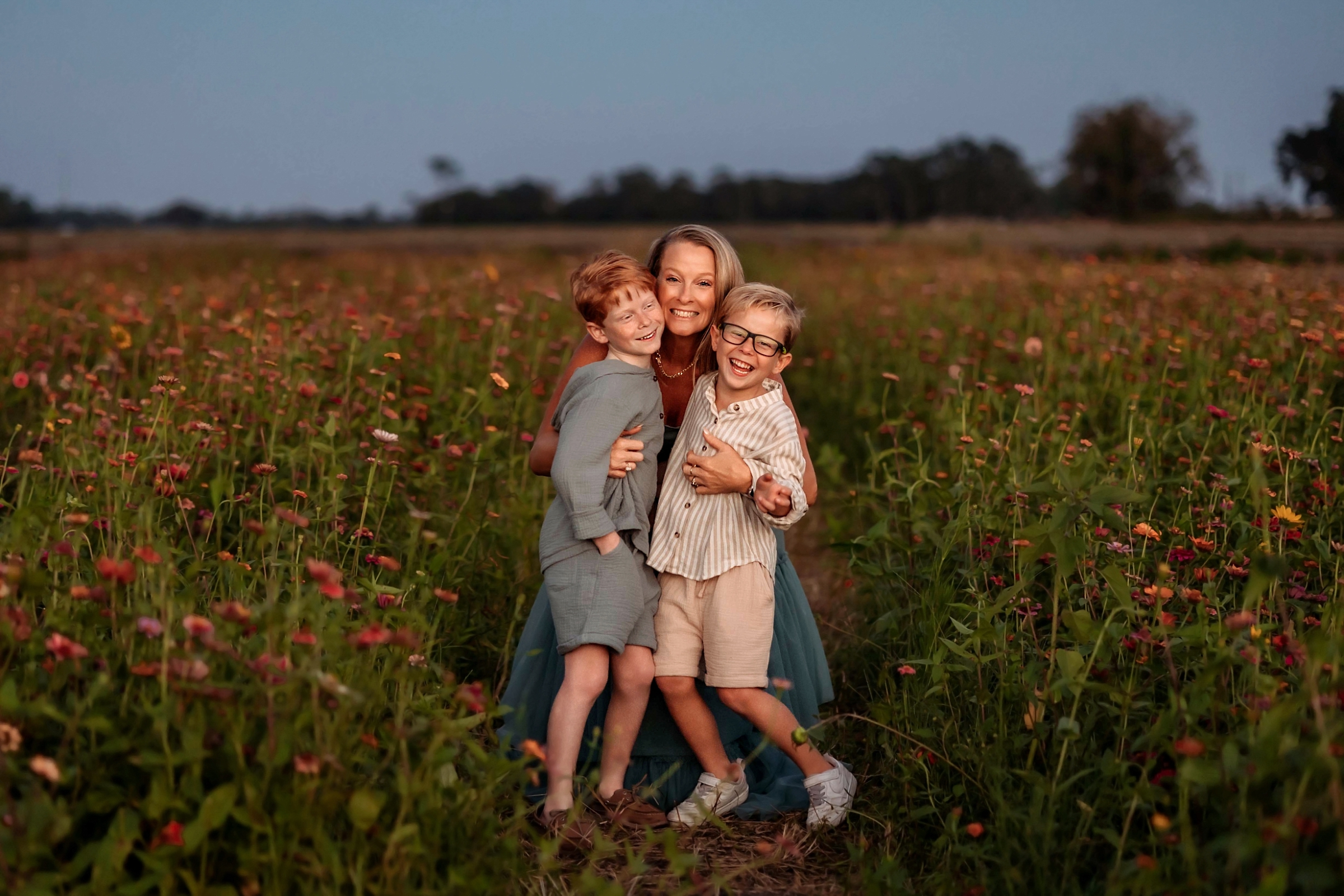 Mother laughing with her two sons in a field of tall wildflowers after sunset.